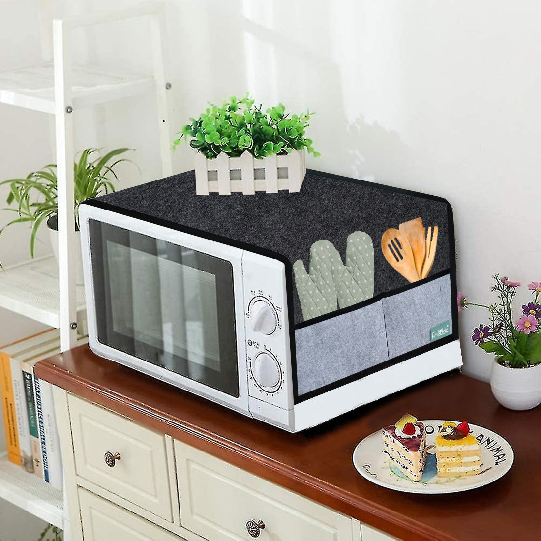 Microwave with a decorative cover on a wooden surface, surrounded by plants and books.