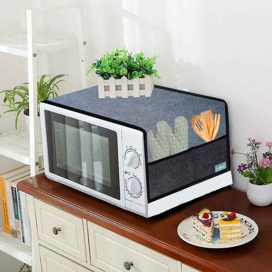 Microwave with a blue cover on a wooden surface, surrounded by plants and books.