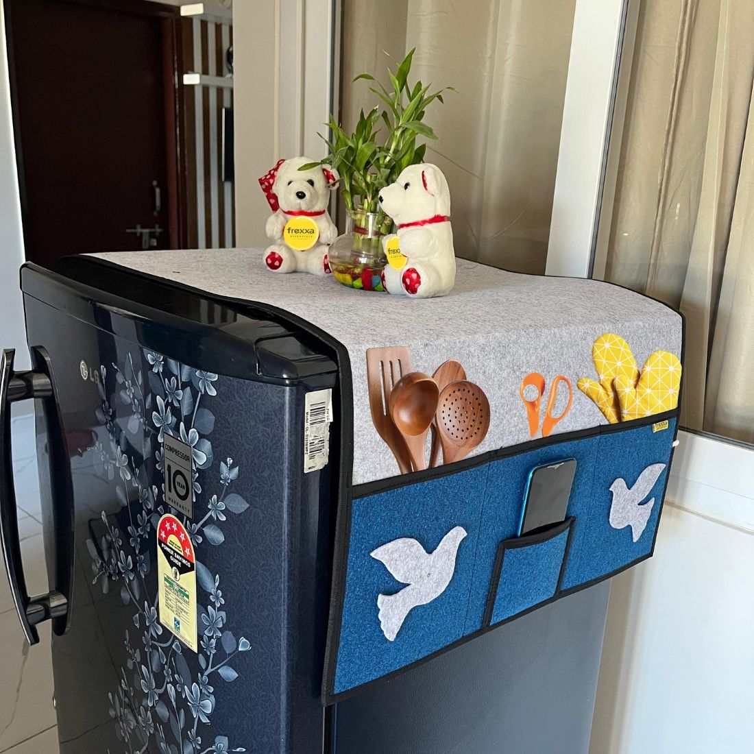 Fridge with a decorative cover featuring utensils and a teddy bear on a countertop.